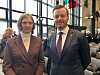 To personer i formelle klær står i en konferansesal med flagg i bakgrunnen