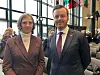 To personer i formelle klær står i en konferansesal med flagg i bakgrunnen