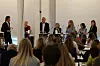 Paneldeltakere står bak høye bord og taler til et sittende publikum i sal.
