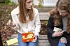To jenter sitter på en benk ute og spiser lunsj fra matbokser.