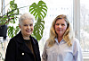 To kvinner poserer sammen i et lyst kontor foran et vindu og en stor grønn plante.