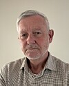 Eldre mann ser alvorlig inn i kamera foran lys, ensfarget vegg.