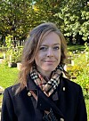 Kvinne med rutete skjerf står i solfylt park med grønt gress og trær bak.