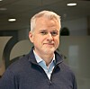 En mann i blå genser står innendørs foran en kontorvegg.