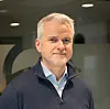 En mann i blå genser står innendørs foran en kontorvegg.