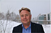 En mann i mørk jakke står ute foran snø og bygninger.