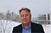 En mann i mørk jakke står ute foran snø og bygninger.