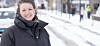Smilende kvinne i mørk vinterjakke står ute i et snødekt bymiljø.