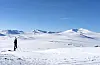 Skiløper går over snødekt vidde med Snøhetta i bakgrunnen under blå himmel.