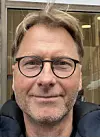 Nærbilde av en mann med briller foran en glassfasade.