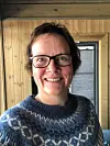 Smilende person med briller i blå mønstret genser foran trevegg og vindu.