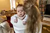 Mor med baby på armen i en stue med sofa, teppe og bokhylle i bakgrunnen.