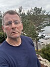 Mann i blå genser står foran trær med småbåter og brygger ved vannet bak seg.