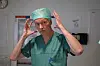 Kirurg i grønn uniform og hette står innendørs med hendene løftet ved hodet.
