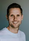 Nærbilde av en smilende mann i hvit T-skjorte foran mørk bakgrunn.