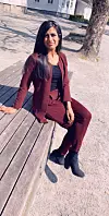 En kvinne i burgunder dress sitter på en trebenk utendørs i sollys.