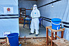 Helsearbeider i vernedrakt står ved inngangen til en midlertidig Ebola-klinikk med blå bøtter og utstyr.