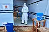 Helsearbeider i vernedrakt står ved inngangen til en midlertidig Ebola-klinikk med blå bøtter og utstyr.