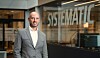 Mann i grå dressjakke står foran en bygning med Systematic-skilt i bakgrunnen.