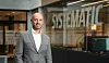 Mann i grå dressjakke står foran en bygning med Systematic-skilt i bakgrunnen.