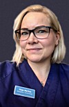 Portrett av en kvinne med briller og blå uniform foran mørk bakgrunn.