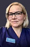 Portrett av en kvinne med briller og blå uniform foran mørk bakgrunn.
