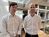 To menn i hvite skjorter står i et lyst kontoratrium med glassvegger.
