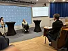 Paneldebatt med deltakere foran en skjerm og publikum i en konferansesal.