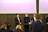 Tre menn står på en scene under en paneldebatt foran et sittende publikum.